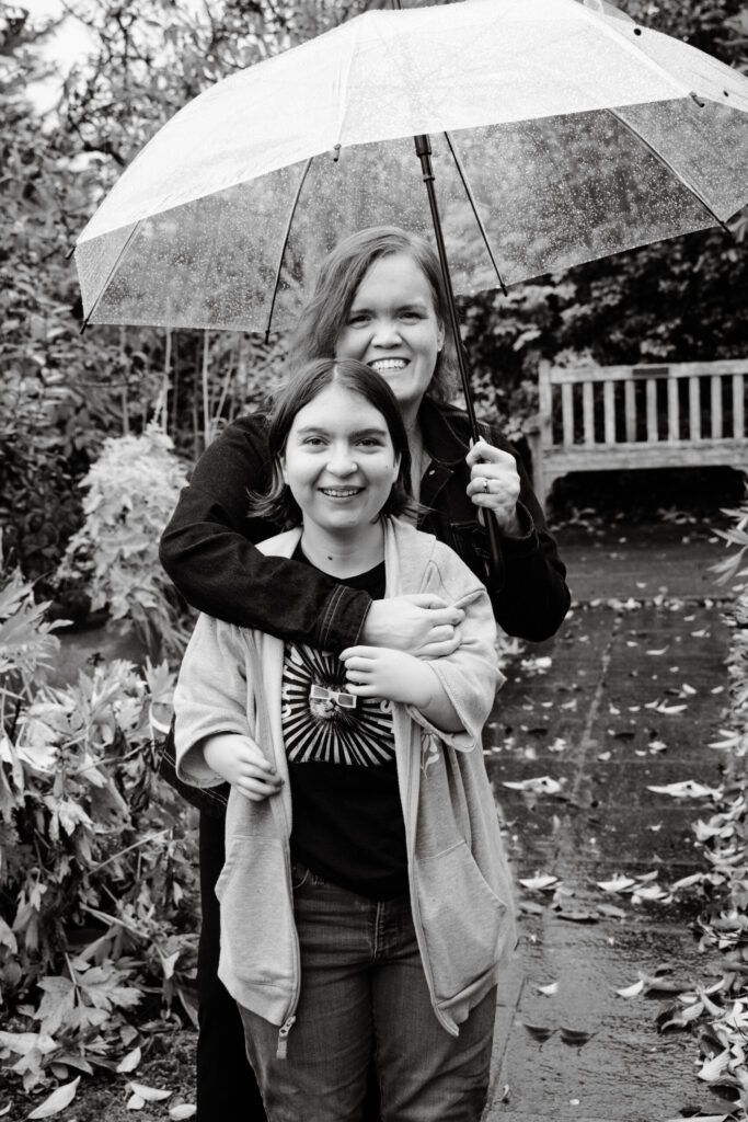 Carrie with her arm around her daughter while holding a clear umbrella